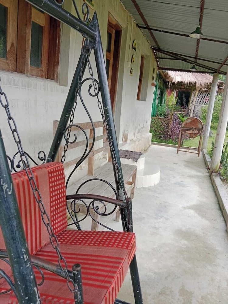 Covered veranda with a red cushioned swing seat and wooden bench, overlooking a garden with a thatched gazebo