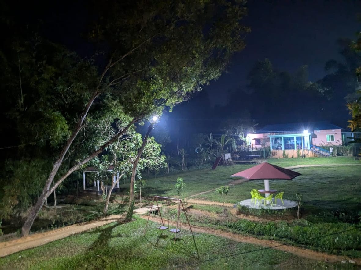 Night view of the resort compound lit up, showing the pink and turquoise cottage, red umbrella-roof gazebo with yellow chairs, and a swing set in the foreground