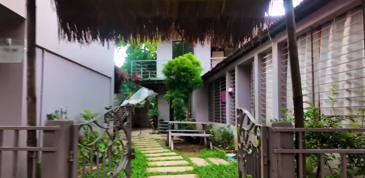 Open wrought-iron gate looking down a stepping-stone path through the guest house courtyard, with a single-storey wing of barred, curtained windows on the right, a thatched awning overhead and a white two-storey block with a first-floor balcony at the end